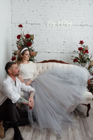 Couple relaxing on vintage sofa against white brick wall with neon sign and red floral arrangements.