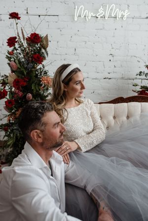 Wedding scene with white brick wall, neon 'Mr & Mrs' sign, and deep red floral arrangements.