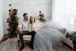 Romantic couple relaxes on white sofa with red roses and greenery decorating bright studio setting.