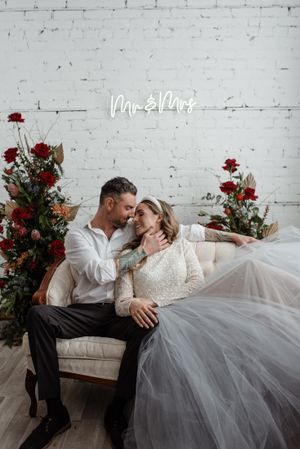 Romantic wedding couple poses on vintage couch with red floral arrangements against white brick wall.