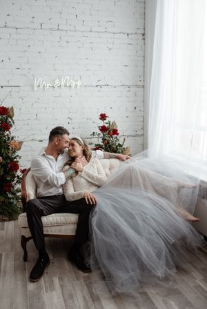 Romantic wedding photo shoot in white studio with tulle dress and red floral decor against brick wall.