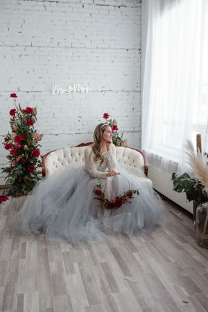 Bride in tulle gown sits on cream couch with red rose arrangements in minimalist white room.