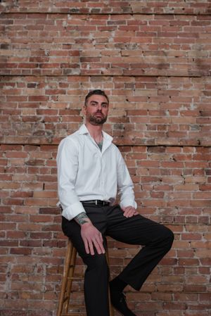 Portrait against brick wall of person in white button-down shirt and dark pants seated on wooden stool.