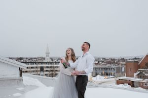 Winter wedding photo with couple joyfully posing in snowy setting with city buildings in background.