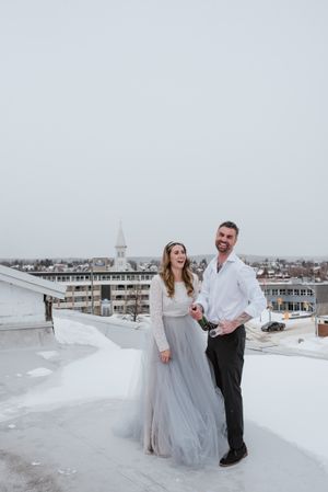 Winter wedding portrait on snowy rooftop with city skyline and church steeple in background.