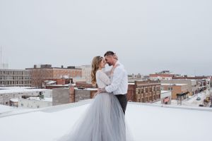 A winter wedding photo on a snowy rooftop with city buildings in the background.