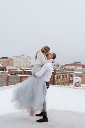 Winter rooftop wedding photo with couple in romantic lift pose against city skyline.
