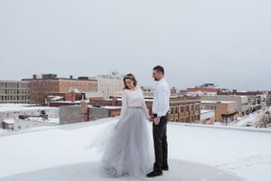 A couple stands together on a snow-covered rooftop with urban buildings in the background.