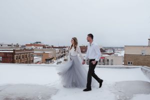 A couple walks hand in hand on a snowy rooftop with urban buildings in the background.