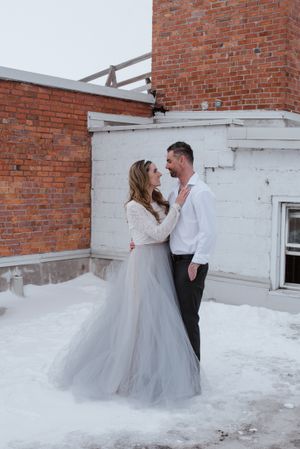 A winter wedding couple embracing on a snowy rooftop with brick walls.