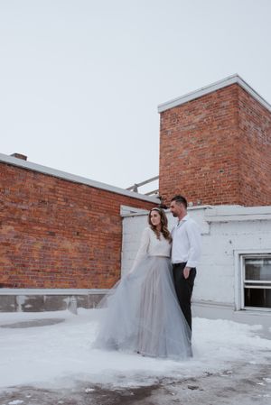 Winter wedding scene with a couple posing against a brick building in the snow.