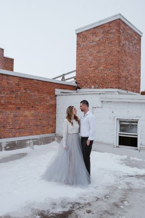 A romantic winter rooftop portrait with snow and brick architecture creating a dramatic backdrop.