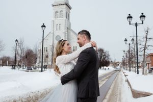 A winter wedding portrait in front of a snowy church with decorative lamp posts.