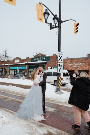 Wedding couple posing on snowy street corner with traffic lights and brick buildings in background.