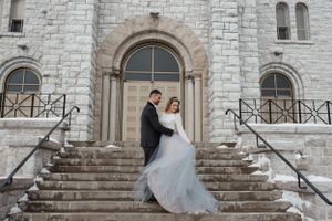 A wedding couple poses on stone steps in front of an elegant church entrance with arched doorway.