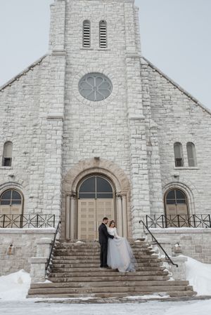 A couple poses on the steps of a white stone church during winter.