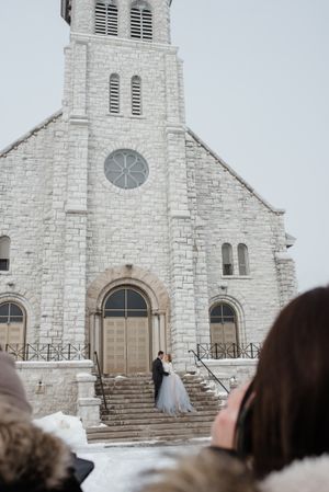 A classic white stone church with couples standing on snowy steps during a winter wedding.
