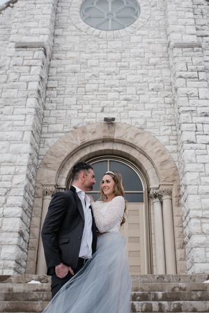 A romantic wedding portrait of a couple in formal attire standing in front of a stone church entrance.