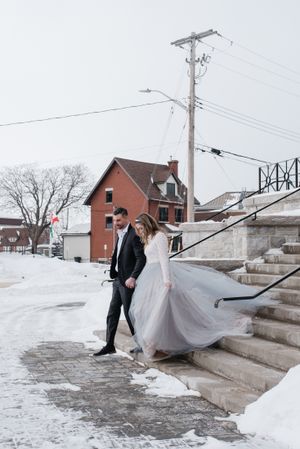 A couple in winter wedding attire walking on snowy steps with a brick building and utility pole in the background.