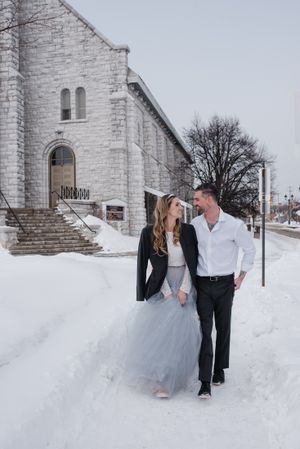 A couple walks together in the snow outside a white stone church.