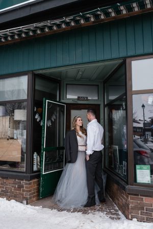 Wedding couple poses in storefront doorway during winter with snow on ground.