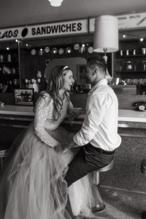 A couple shares a romantic moment at a sandwich shop counter, laughing together in formal attire.