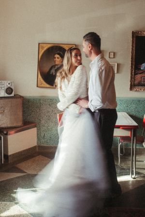 A couple in wedding attire shares a tender moment in a retro-styled room with vintage portraits.