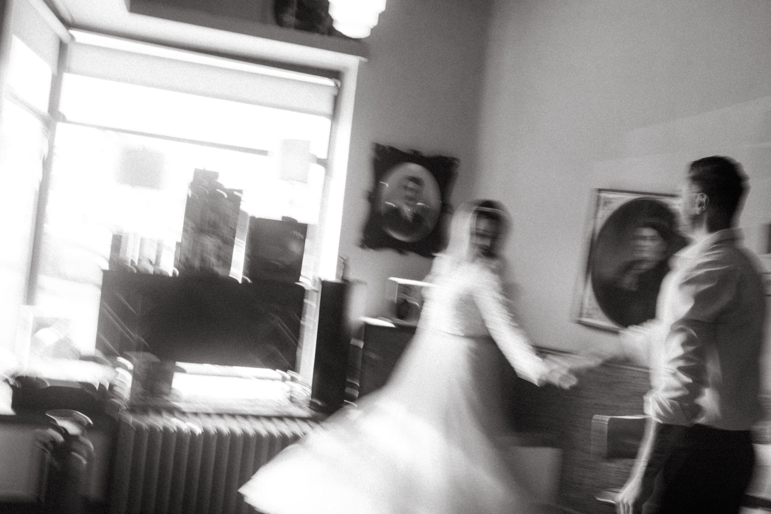A blurred black and white motion shot of two people dancing in a room with portraits on the wall.