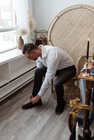 A person in white shirt and dark pants sits on a wicker chair while putting on shoes.