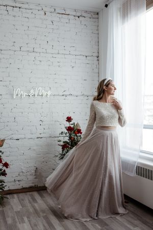 Bride in two-piece wedding dress poses against white brick wall with neon Mr & Mrs sign.