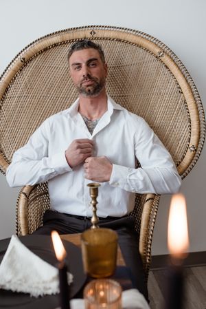 Groom in white shirt seated in wicker peacock chair with candlelit setting.