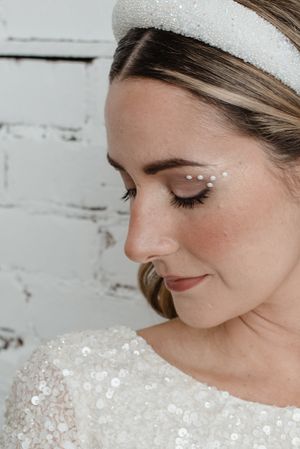 Close-up detail of bridal makeup with pearl accents and a sparkly headpiece against a white brick wall.