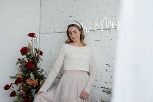 Bride in a two-piece wedding dress posing against white brick wall with neon signage and red flowers.