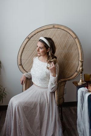 A bride in a white lace dress with long sleeves poses elegantly in a peacock wicker chair against a gray wall.