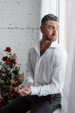 Portrait of a person in white shirt with tattoos looking pensively out a window near red roses.