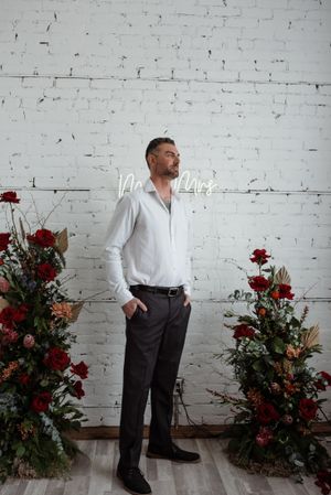 A groom in a white shirt and dark pants poses against a white brick wall between floral arrangements with red roses.