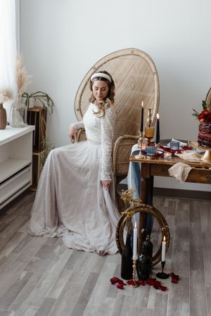 Bride in long-sleeved lace wedding dress sits elegantly in rattan peacock chair with vintage decor and candles nearby.