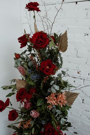 A stunning floral arrangement with deep red roses, eucalyptus, and copper-colored leaves against white brick.