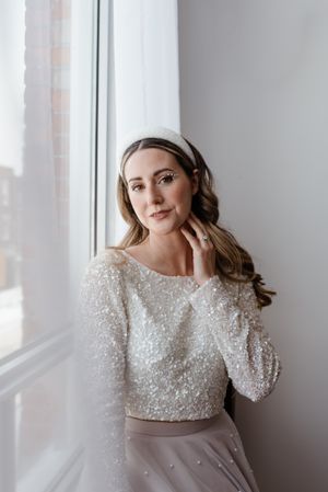 Wedding attire featuring a sparkly long-sleeved crop top and tulle skirt in soft gray tones by a window.