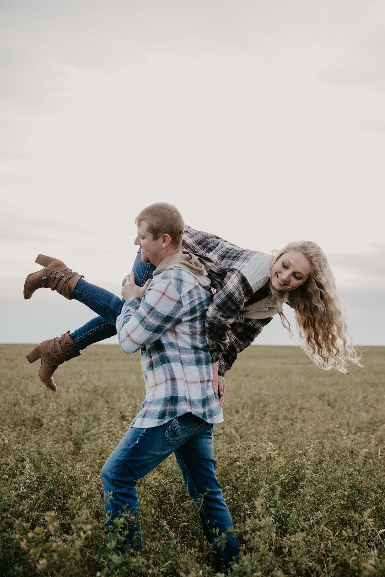 couple laughing as he carries over his shoulder through the tall grass as the sun sets behind them