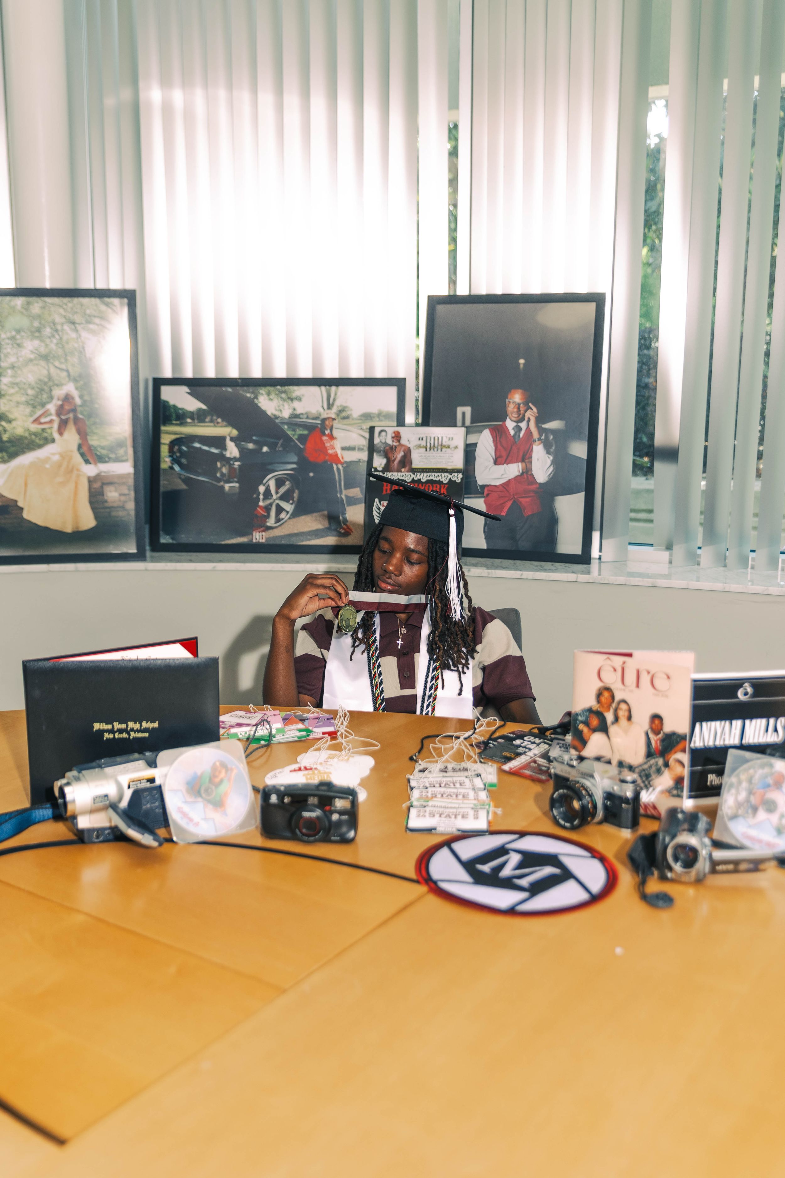 Mills in graduate in cap and gown sitting at desk surrounded by cameras, magazines, and framed photos.