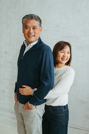 A couple wearing blue and white casual outfits embrace and smile together against a white brick wall.
