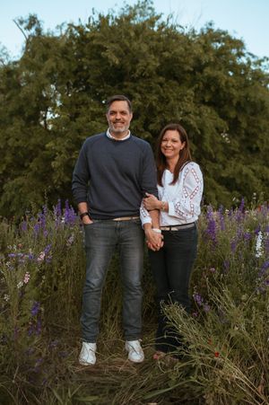 A couple stands together in casual attire among purple wildflowers and green foliage during a sunset photo session.