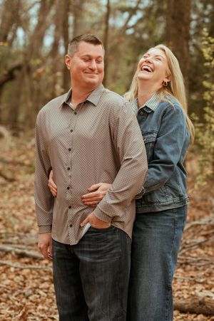 Two people in autumn clothing pose playfully on fallen leaves in a forest during golden hour sunset.