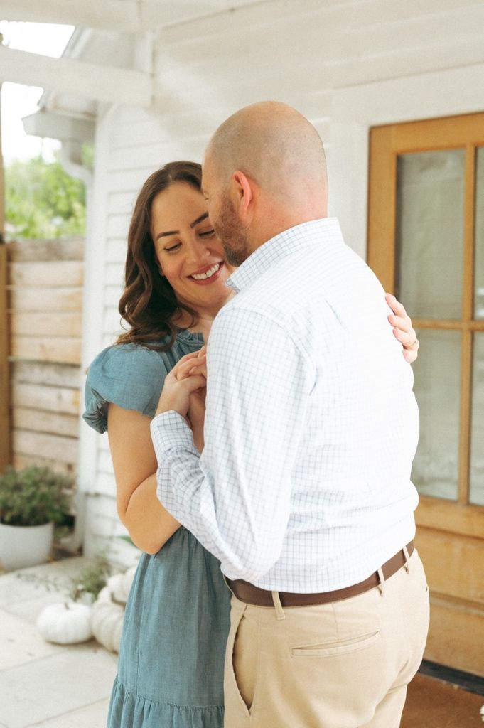A smiling woman in a teal dress embraces a bald man in a light blue shirt on a charming white porch.