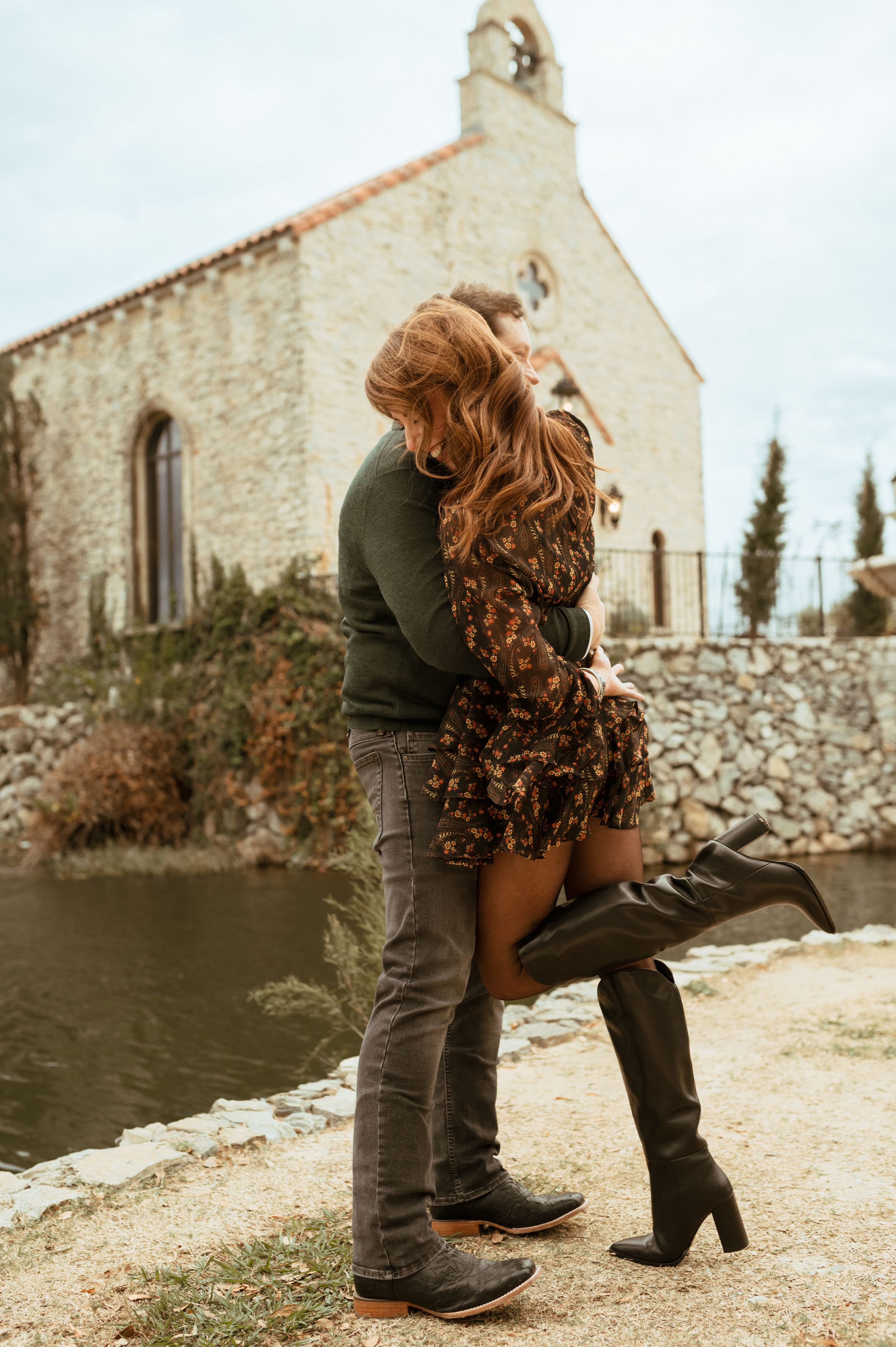 A romantic couple embraces near a stone church by the water on an autumn day.