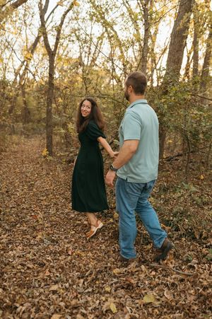 A couple playfully runs through fallen autumn leaves in a wooded area during a photo session.
