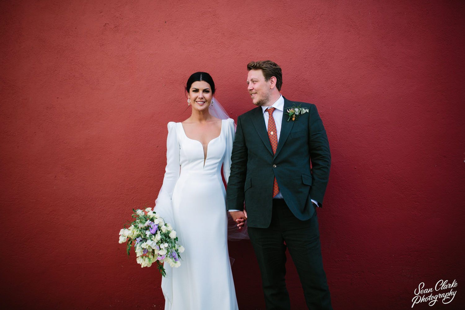 Wedding couple stands against vibrant red wall, bride in long sleeve white gown holding floral bouquet.