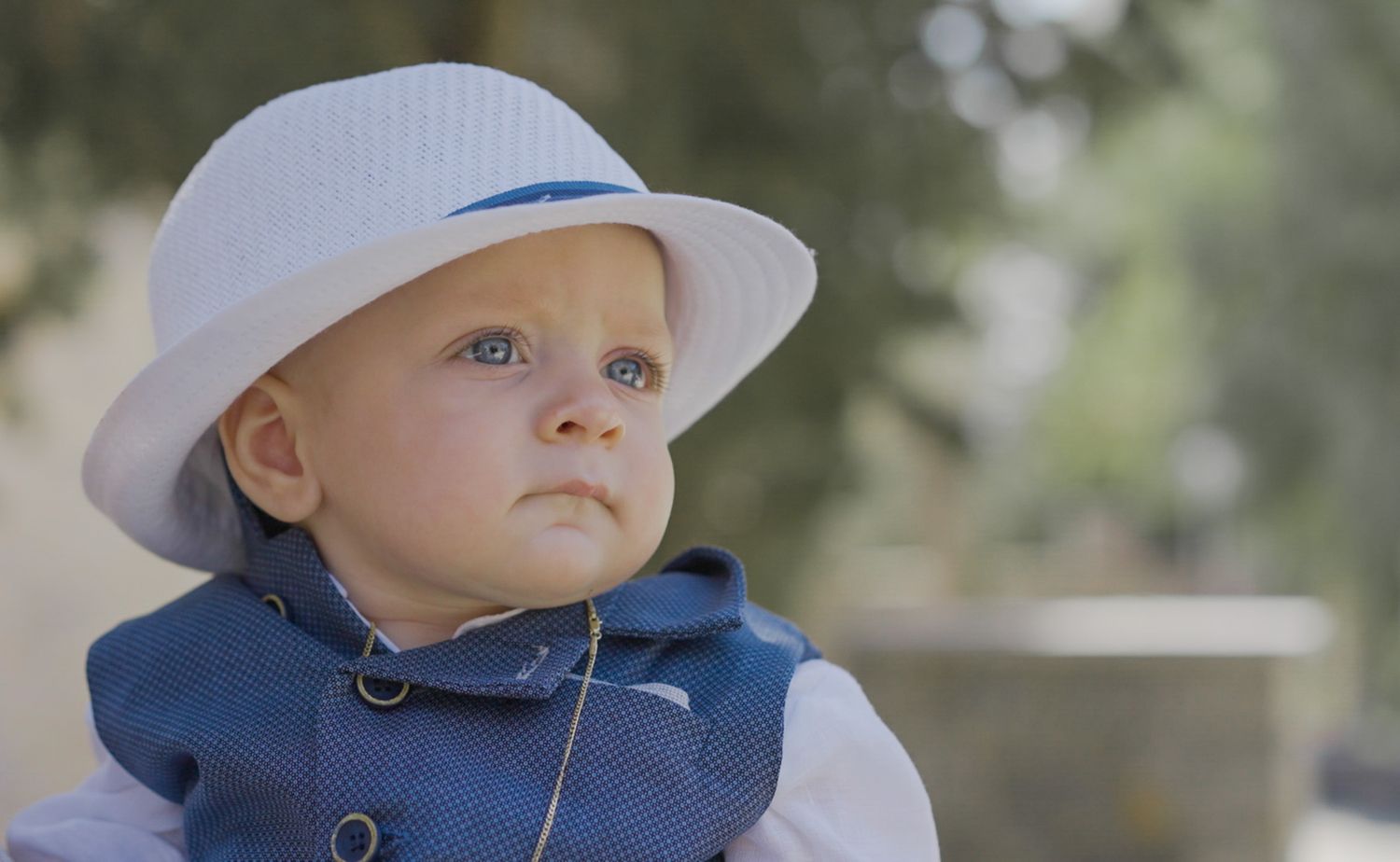 A small child wearing a white sun hat and navy jacket gazes upward thoughtfully.