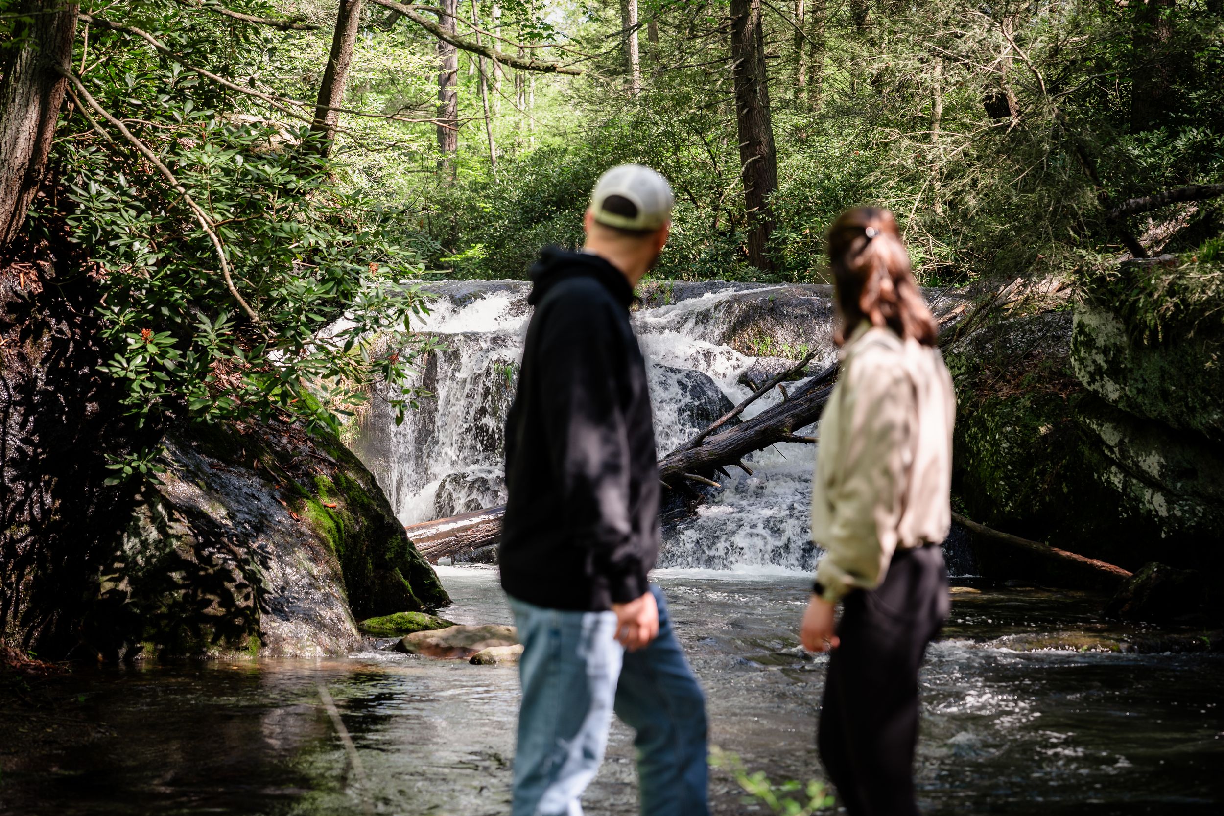Couple walking along a wooded path near a flowing stream surrounded by green foliage.